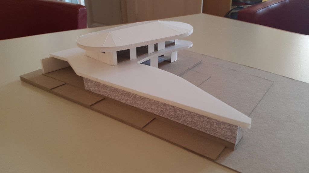 Foto eines physischen Architekturmodells aus weißer Pappe auf einer grauen Grundplatte, das ein modernes Gebäude mit geschwungenem Dach und einer gemauerten Sockelwand darstellt.