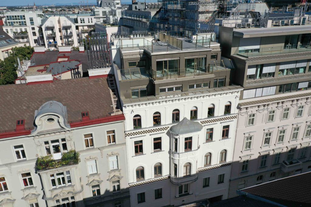 Vogelperspektive auf ein klassisches Altbaugebäude mit verzierter weißer Fassade und einem modernen, mehrstöckigen Dachausbau samt Terrassen in einer dichten städtischen Häuserzeile.