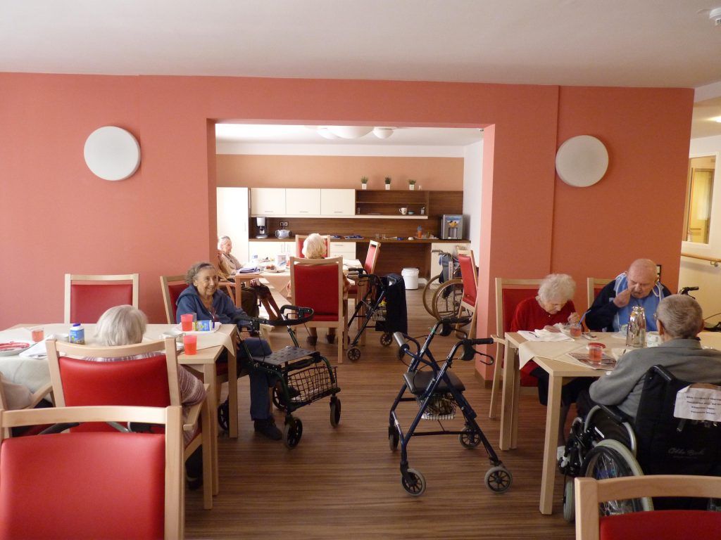 Gemeinschaftsraum einer Senioreneinrichtung oder eines Pflegeheims mit älteren Menschen an Esstischen, Rollatoren und einer Küche im Hintergrund.