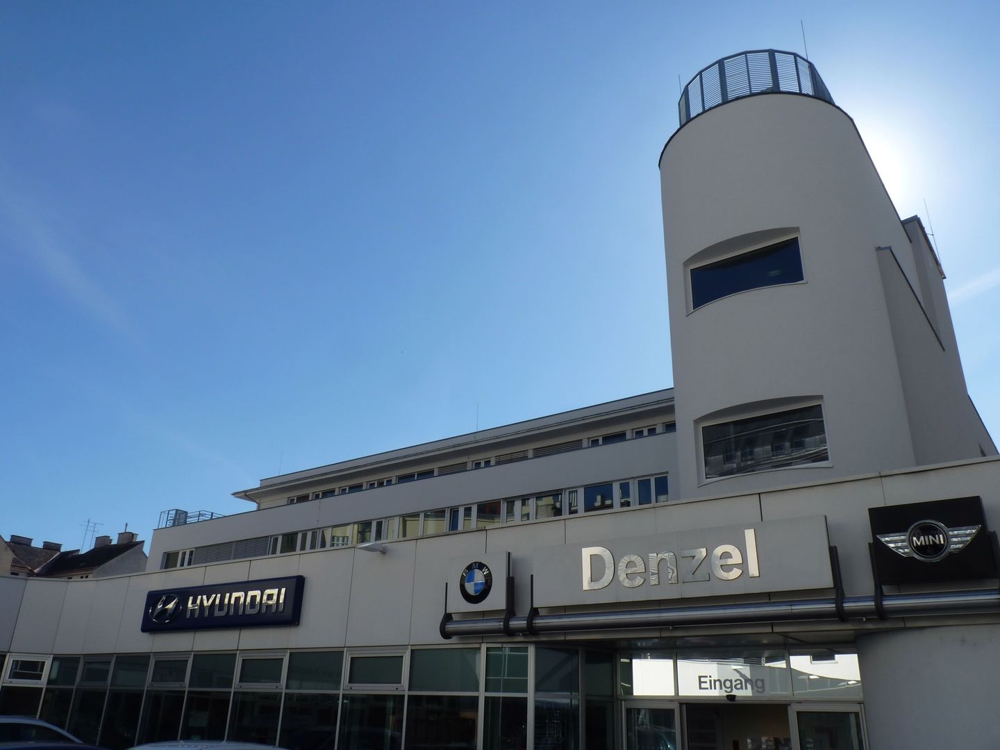 Moderne, weiße Fassade des Autohauses Denzel mit Logos von BMW, MINI und Hyundai unter blauem Himmel.