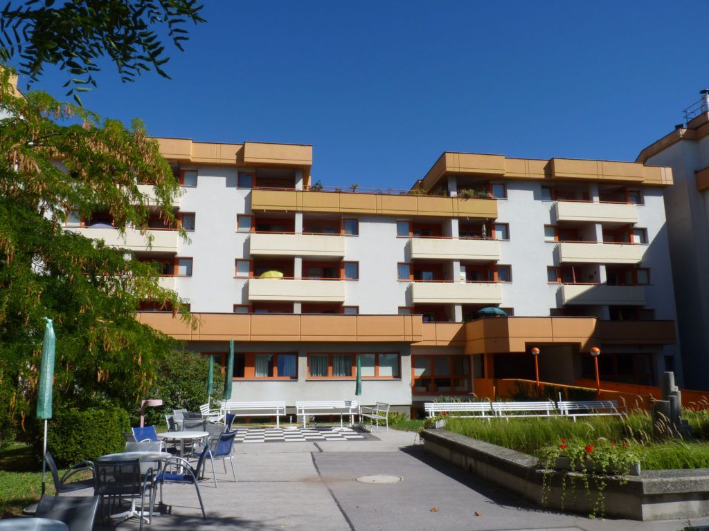 Mehrstöckiges Wohngebäude mit Balkonen, weiß-oranger Fassade und einem gestalteten Innenhof inklusive Außensitzplätzen unter blauem Himmel.