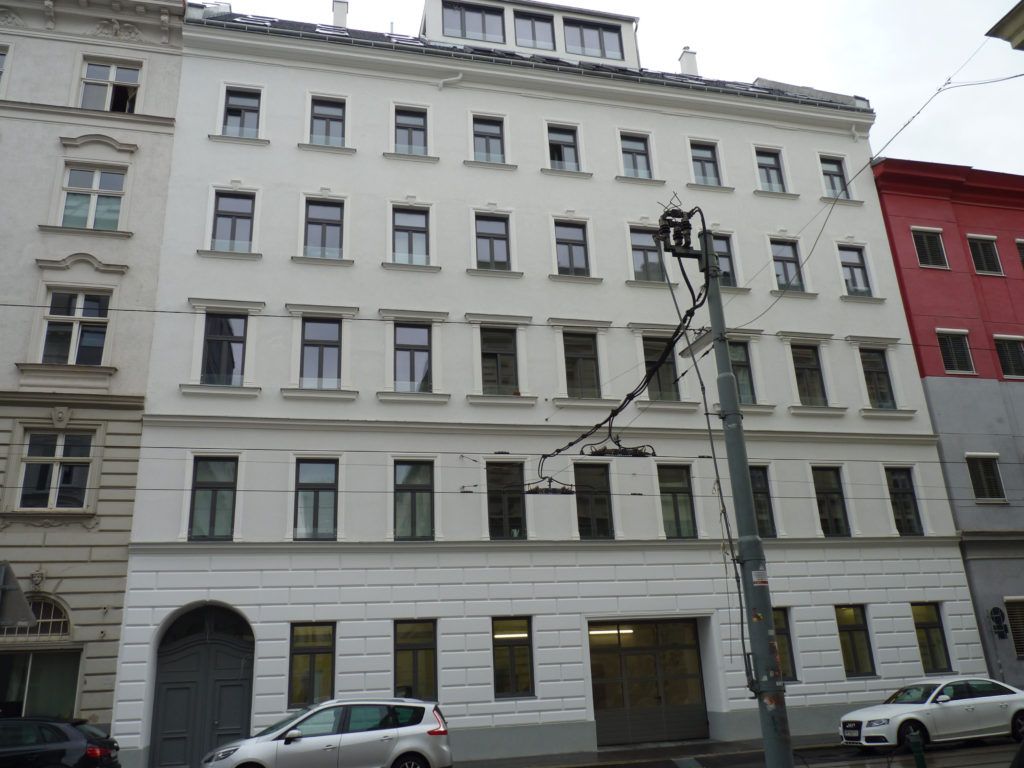Frontalansicht eines mehrstöckigen, weiß gestrichenen Stadthauses mit symmetrischer Fensteranordnung, einem großen Einfahrtstor und parkenden Autos an der Straße.