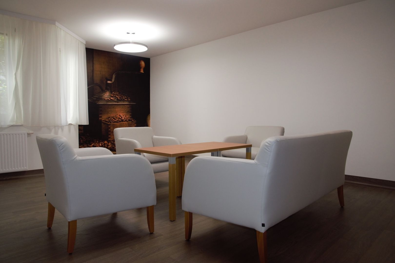 Innenansicht eines Besprechungs- oder Warteraums mit einer Sitzecke bestehend aus einem Sofa, drei Sesseln, einem Holztisch und einer Bildtapete.