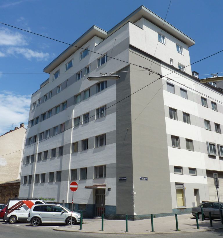 Außenansicht eines mehrstöckigen Wohngebäudes an einer Straßenecke mit einer weiß-grau gestreiften Fassade unter blauem Himmel.